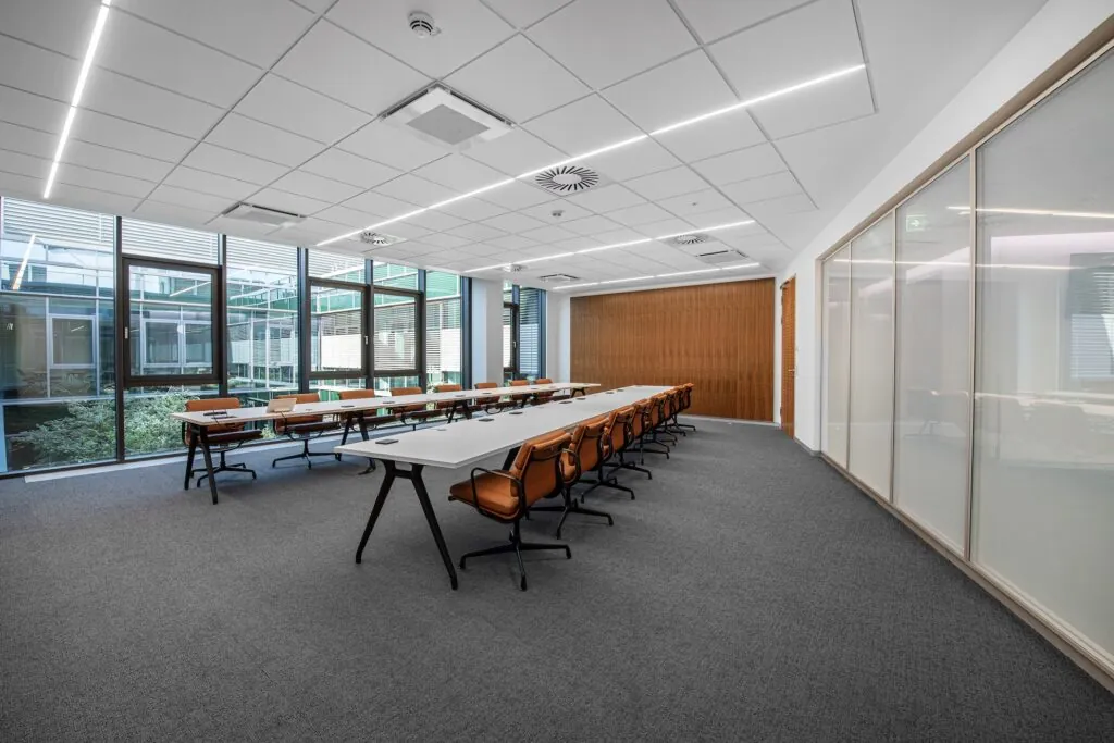 Meeting room with long tables, chairs, carpet flooring and large window surfaces in a modern office building.